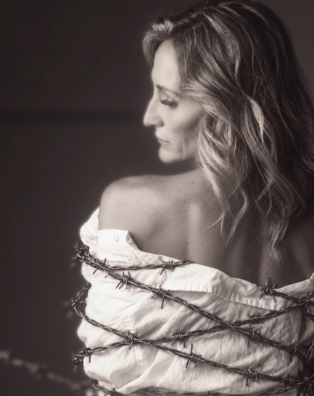 Sepia portrait of Jenn Board looking over her bare shoulder, a white shirt wrapped tightly in barbed wire around her body