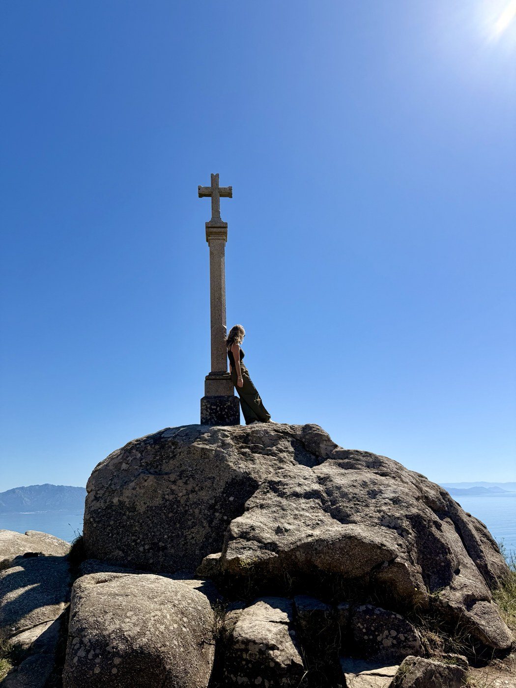 Jenn Board leaning against the stone cross at Finisterre, Spain, at the end of her Camino, with the ocean and mountains behind her.